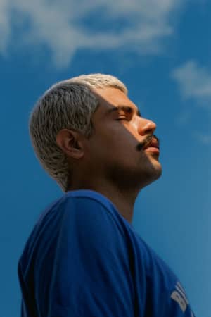 A young man with bleached hair and a dark moustache raises his head to the sun with a blue sky and clouds behind him.