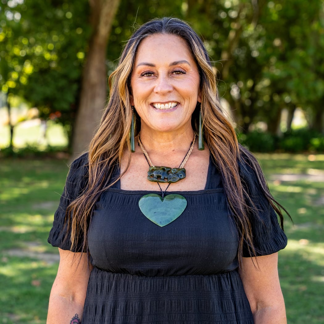 Kahira smiles at the camera. She is standing outside wearing a black top and a large taonga around her neck - a tiki, and a large pounamu heart.