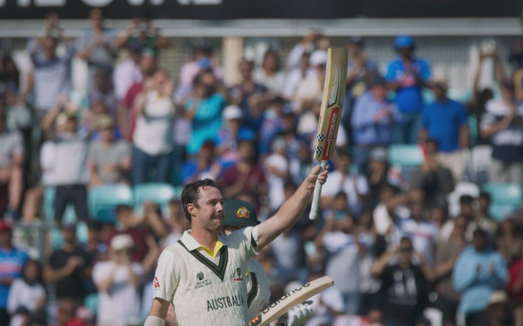 Australian cricketer Travis Head holds his bat in the air with a crowd of spectators in the background.
