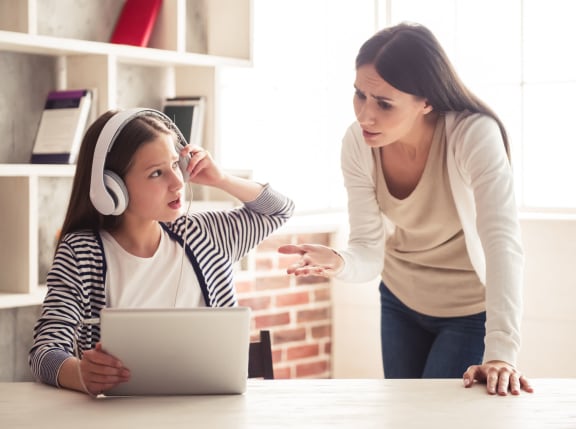 A photo of a troubled teenage girl in headphones using a digital tablet while her mom is scolding her