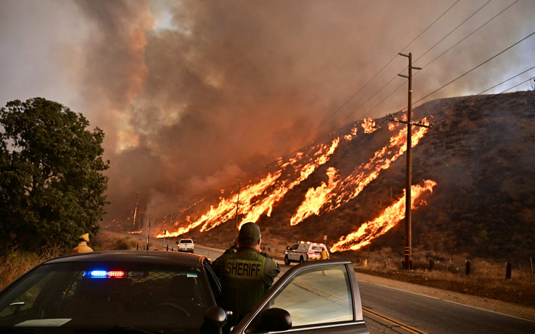 A sheriff watches flames race up the hill as the plume of smoke from the Hughes Fire fills the sky in Castaic, a northwestern neighborhood of Los Angeles, California, on January 22, 2025. A new wildfire erupted north of Los Angeles on January 22, exploding in size and sparking thousands of evacuation orders in a region already staggering from the effects of huge blazes.
Ferocious flames were devouring hillsides near Castaic Lake, spreading rapidly to cover 5,000 acres (2,000 hectares) in just over two hours.
The fire was being fanned by strong, dry Santa Ana winds that were racing through the area, pushing a vast pall of smoke and embers ahead of the flames.
Evacuations were ordered for 19,000 people all around the lake, which sits around 35 miles north of Los Angeles, and close to the city of Santa Clarita. (Photo by Frederic J. Brown / AFP)