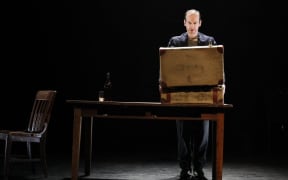 Denis O'Hare stands on a dark stage in front of a suitcase on a table.