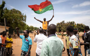 A man waves a Burkina Faso flag as others demonstrate while Burkina Faso soldiers are seen deployed in Ouagadougou on September 30, 2022. - Shots rang out before dawn on Friday around Burkina Faso's presidential palace and headquarters of the military junta, which itself seized power in a coup last January. The government said the developing situation was linked to an "internal crisis in the army", after AFP journalists saw troops block several main roads in the capital Ouagadougou. (Photo by Olympia DE MAISMONT / AFP)