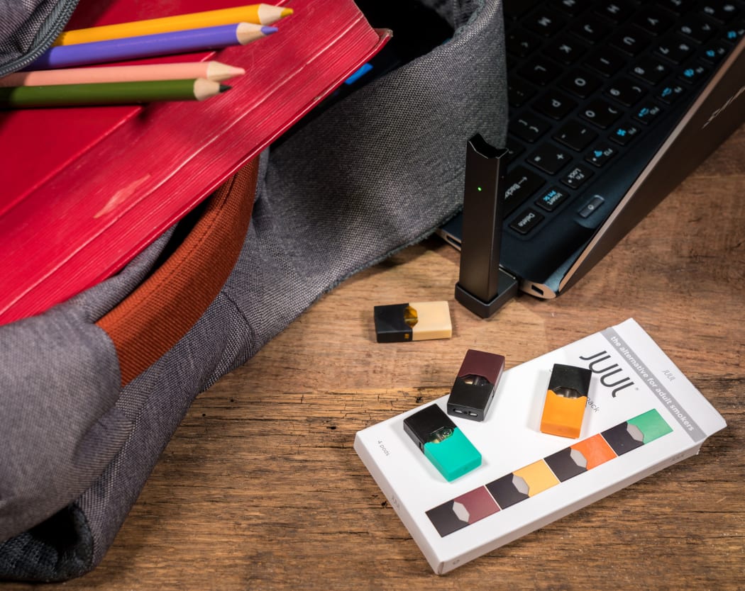 A JUUL device charges on a laptop, beside JUUL pods.