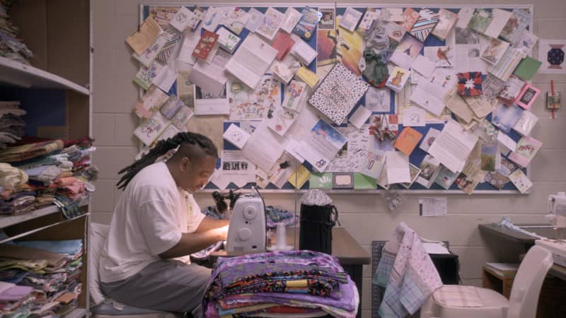Chill in Netflix documentary, The Quilters, directed by Jennifer McShane, sits at a sewing machine table near a wall filled with cards and notes.