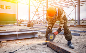 A strong  man welder in brown uniform, a construction helmet and welders leathers, grinder metal an angle grinder  at the construction site, orange sparks fly to the sides