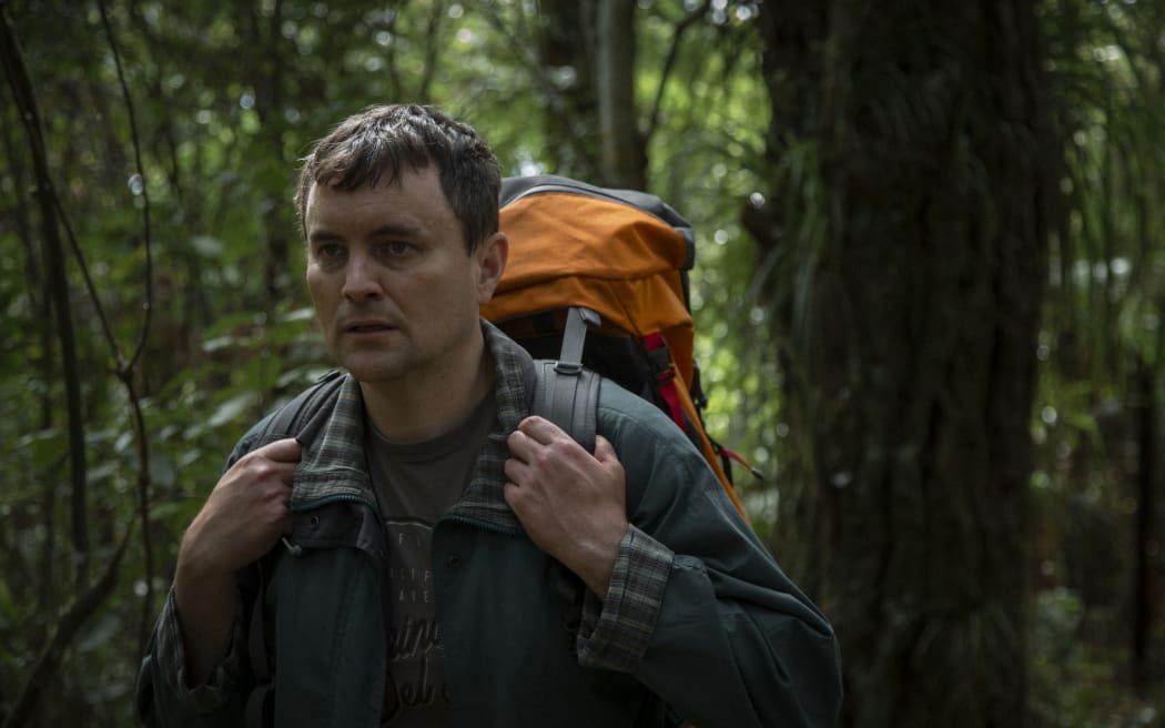 Stressed-looking man in his early 40s alone in the New Zealand bush carrying a tramping pack.