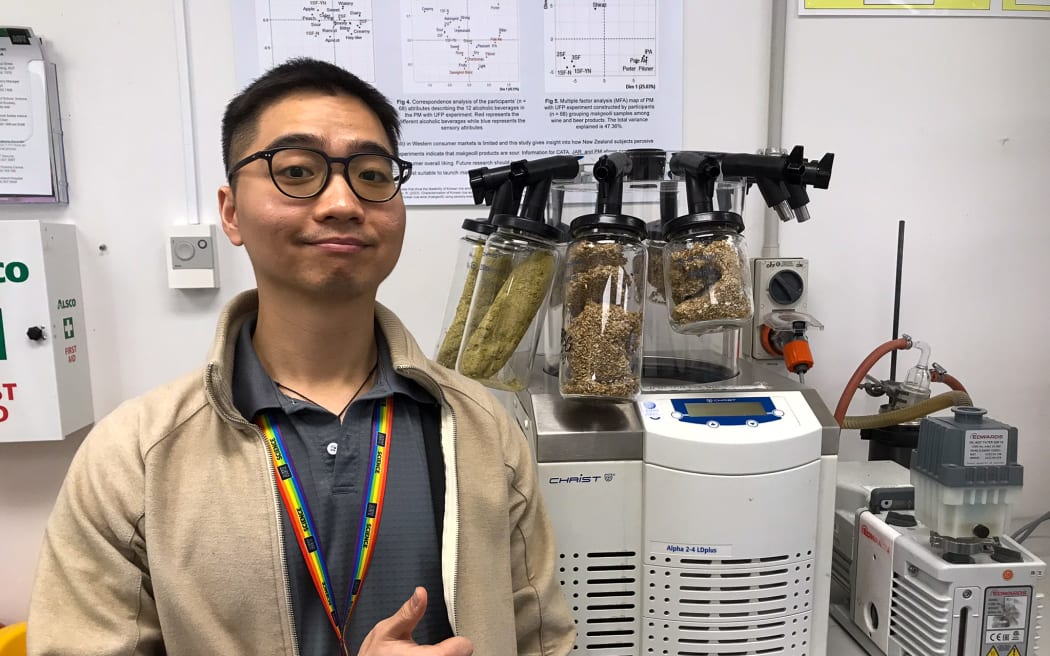 A man wearing glasses, a beige sweater and a rainbow lanyard stands in front of a piece of lab equipment with various different grains in plastic containers attached to it. He is smiling and giving a "thumbs up".