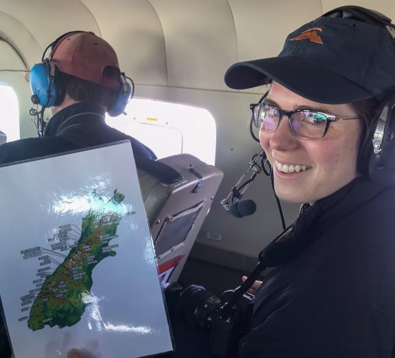 A woman wearing headphones and headset, a cap, and glasses is sitting in a small plane. She is smiling at the camera while holding up a laminated map of the South Island.