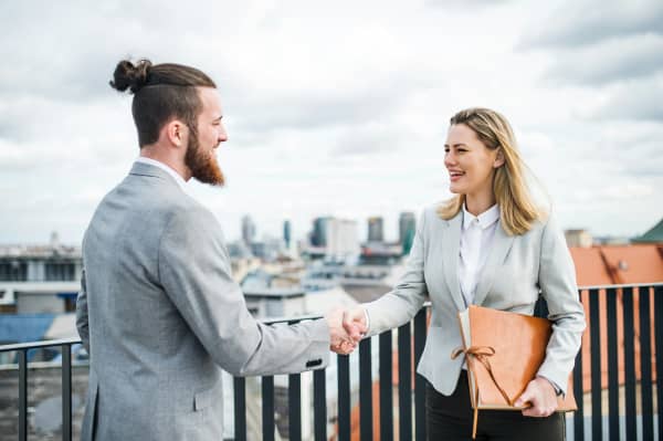 A man and woman in grey suit jackets shake hands on an outdoor balcony.