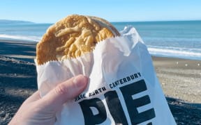 A person holds a pie up in front of a beach.