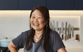 Nagi stands in her kitchen, smiling, wearing a grey apron. Behind her on the kitchen bench is a crockpot, and an assortment of chef's knives.