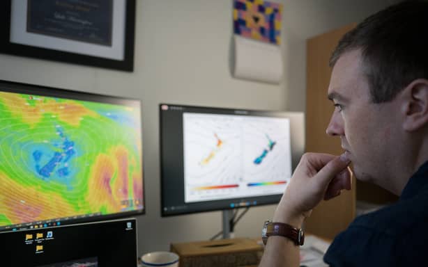 A close up shot of Luke looking at two monitors which have weather and climate maps of New Zealand up on them. He has his thumb and a finger to his lips, as if concentrating.