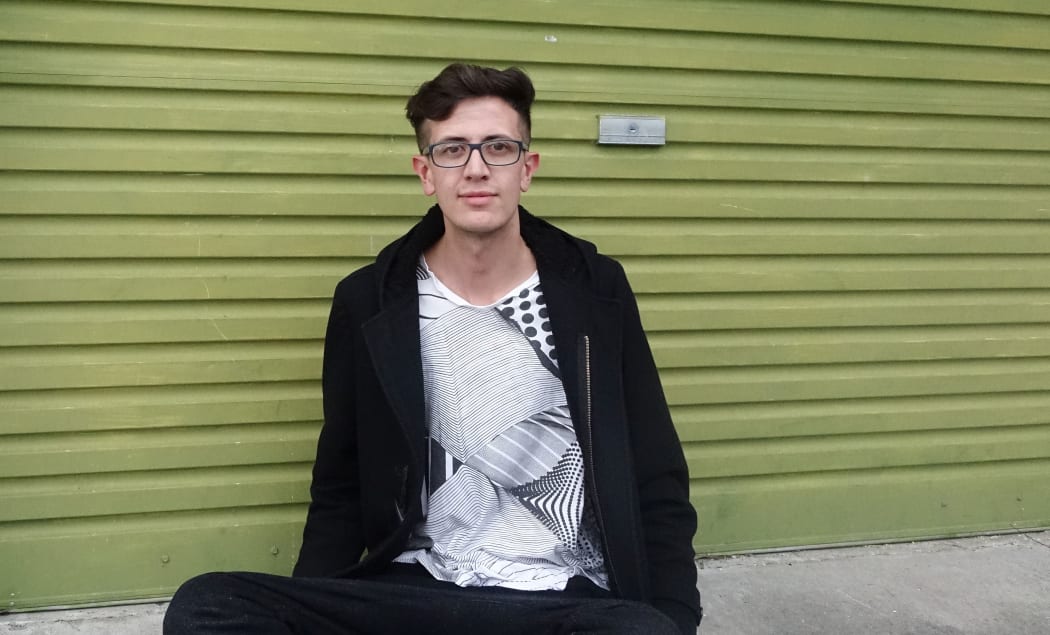 Christian who is in his 20s sits in front of a green painted metal garage door