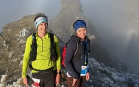 Two people in outdoors gear wearing backpacks are smiling, standing atop a craggy mountain peak shrouded in mist.