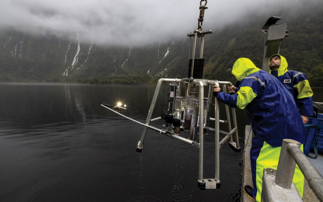 Two men dressed in bright yellow and blue boat overalls are steadying a shiny metal piece of equipment that looks like a moon lander as it is lowered beside the boat. In the background you can see a fiord landscape - lush mountains, dark sea, low cloud and waterfalls.