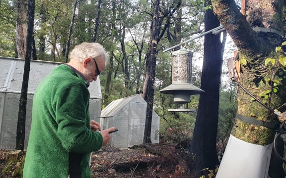 Kevin is wearing a green fleece and sunglasses. In the photo he is looking down at his phone, logging something. He is standing in front of a large bird feeder with some mesh sheds in the background, in the forest.