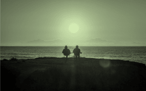 The image is of a pair of people in the foreground on a beach, watching the sun go down. Both are sitting on folding chairs.