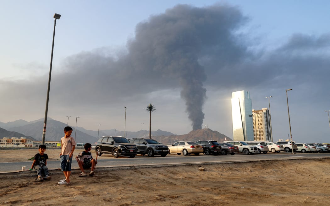 Boys sitting on the side of a road watch as behind a tall smoke plume billows following an explosion in the Fujairah industrial zone on March 3, 2026. Iran's strikes on Gulf neighbours since February 28, following the US-Israeli attack, forced the UAE to shut its airspace, blindsiding travellers who thought they were headed to one of the region's safest holiday destinations.