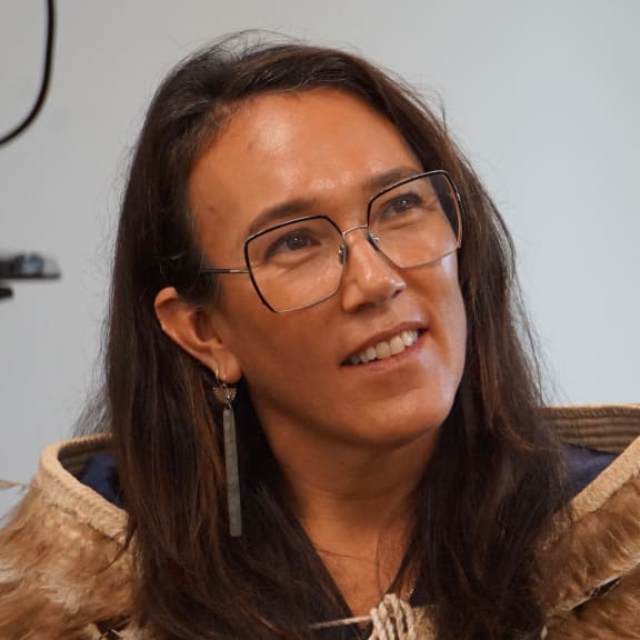 Claire Charters is wearing glasses with long brown hair, she is sitting down and is looking upwards, as if she is listening.