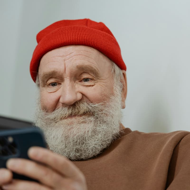 A senior man with a beard and red beanie smiles at his phone.