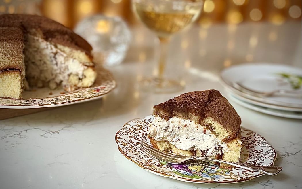 A whole Zuccotto (a dome of sponge filled with whipped cream laced with chocolate and hazelnuts) sits on a plate with a wedge cut out of it, the wedge is on a plate in the right foreground. There is a glass of prosecco and fairy lights in the background.