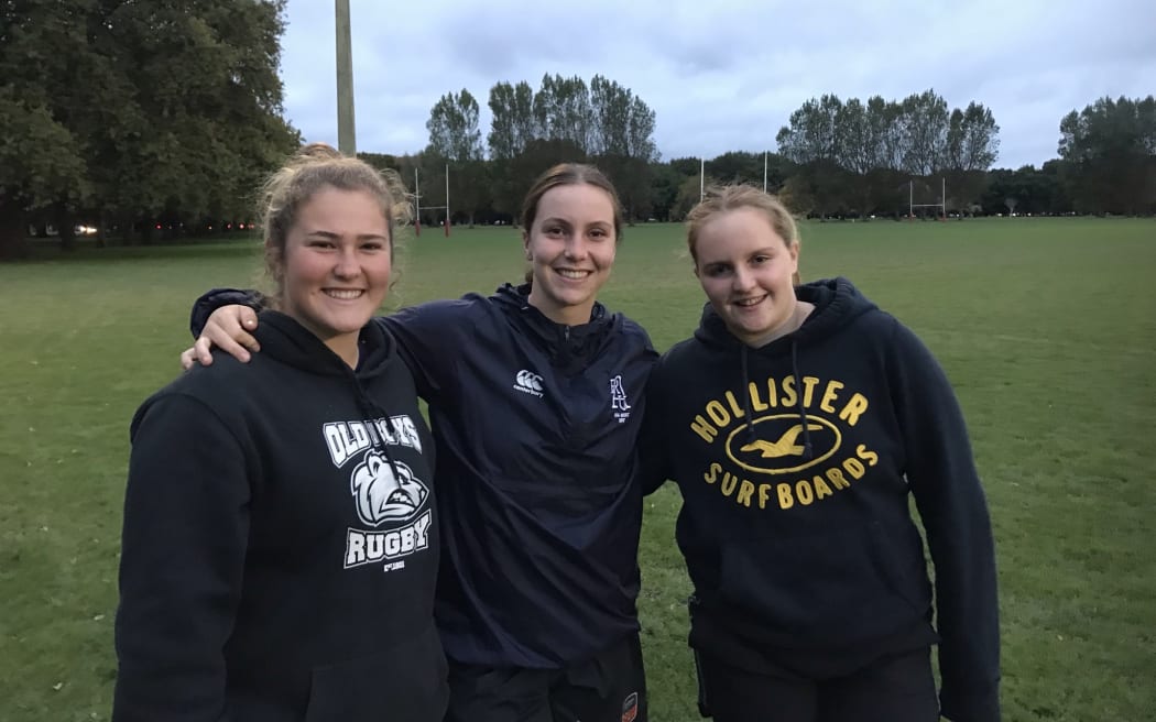 Three teenage girls are standing on a sports playing field with their arms around each other, facing the camera and smiling. They're wearing hoodies.
