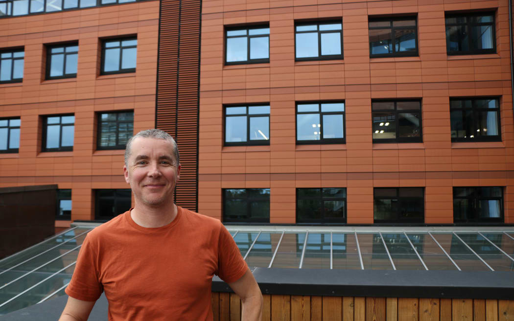A midshot of man in a burnt orange t-shirt standing on a balcony in front of a brick building. He is smiling at the camera.