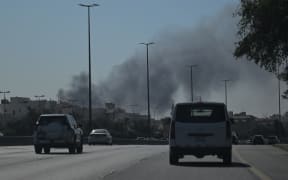 Motorists drive along a street as smoke rises from a reported Iranian strike in the area where the US Embassy is located in Kuwait City on March 2, 2026. Black smoke was seen rising from the US embassy in Kuwait City on March 2 after the latest volley of Iranian strikes, an AFP correspondent saw. (Photo by AFP)