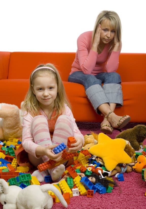 917736 - 5-6yo girl with her toys on the floor. there's huge mess. there's mother sitting on couch behind girl. mother has headache cause this mess.