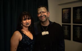 A man and a woman together, one has a full face moko kauae and the other has a moko kauae on her chin.