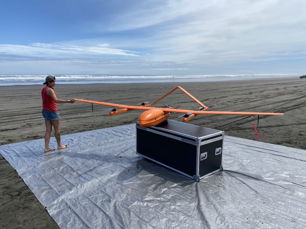 Hayley Nessia with the Maui63 project drone before launch. The large orange drone is on a big case on top of a large sheet of grey plastic on a beach.