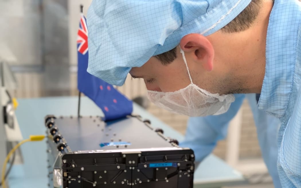 Ben Taylor is wearing a blue clean suit, including a hair net and facemask. He is building a satelite on a lab bench. The satelite has a New Zealand flag on it.