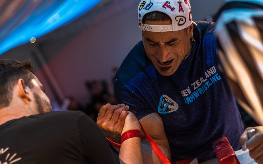John Henry Leach is armwrestling. He is wearing a white baseball cap and blue "New Zealand Armwrestling" federation tshirt. The cap is on backwards. He has a look of intense concentration on his face.