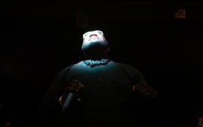 Maga Ran standing in the shadows, his face turned up wards with a spotlight shining on it, wearing a tie that says Hustle.