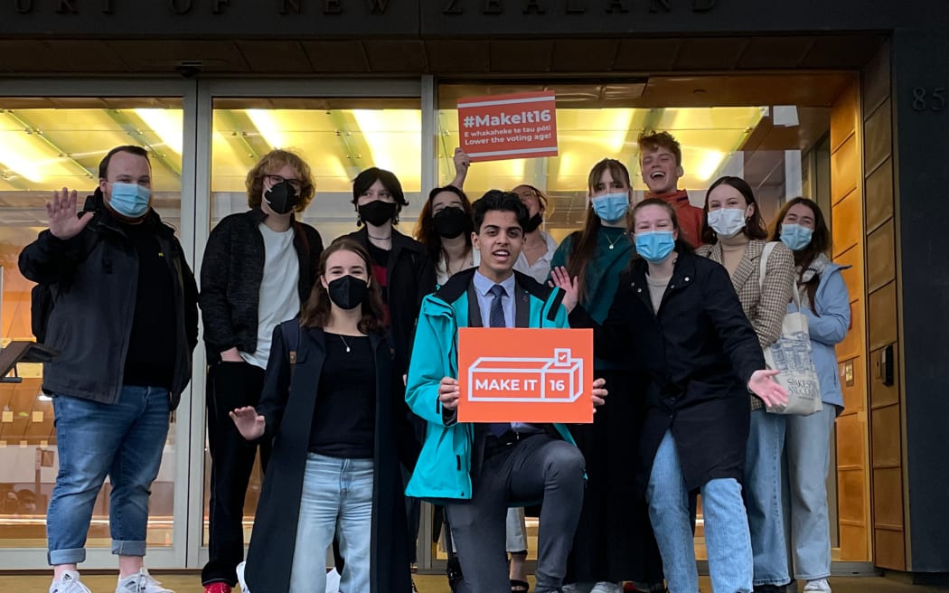Group of young people outside court building in Auckland, one holding an orange placard that reads 'Make it 16'