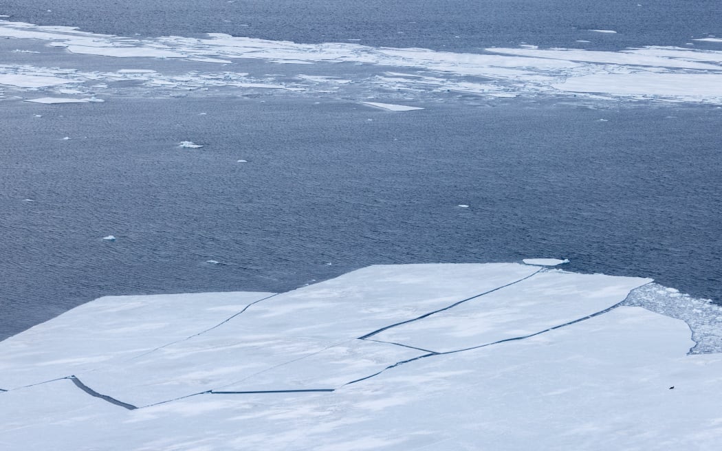 A wide shot of ice floes breaking apart, as seen from a high angle. Beyond the ice edge, there is open dark water, then a line of mushy ice.