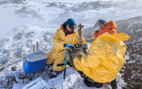 Two people in yellow hazmat suits wearing beanies and eye goggles kneel in the snow and dark volcanic rock of a mountain slope. Volcanic steam makes the scene below hazy: black outcrops peeking out of smooth snow and ice. They are fixated on a frame contraption sitting on the snow between them, with two chilly bins resting nearby.