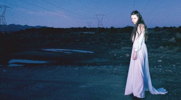 Greta O'Leary - a white woman with long brown hair wearing a white dress - stands in an industrial landscape at dusk.