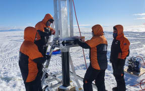 Four scientists in Antarctica, wearing orange and black jackets. They are standing around a piece of silver equipment.