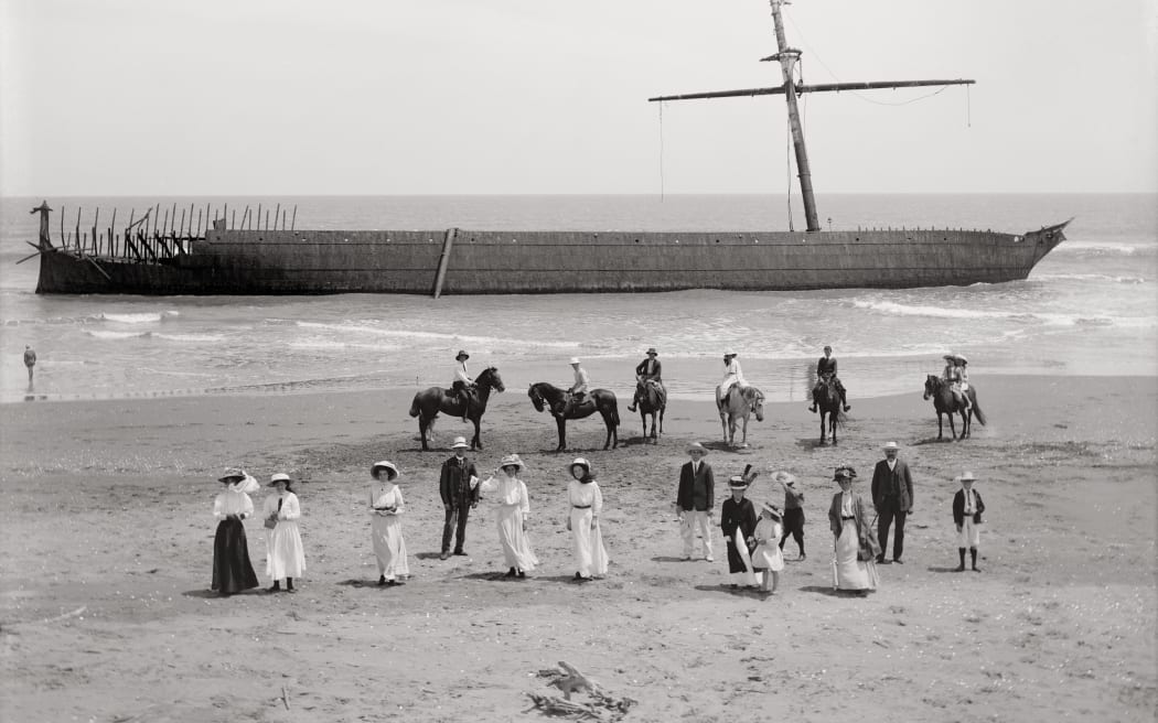 A group of people and some horses gathered on the beach. A large ornate boat stands in the background, stopped in shallow shores.