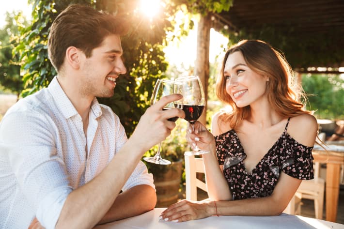 Picture of young loving couple sitting in cafe by dating outdors in park holding glasses of wine drinking.