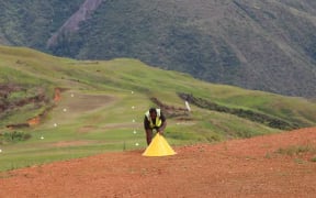 A Papua New Guinea Rural Airstrip Agency Maintenance Officer in Sopu, Goilala places a cone marker. The airstrip and windsocks are visible in the background and mark the taxi way in the distance.
