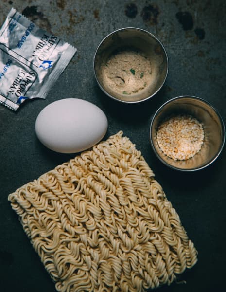 Ingredients on a table; an egg, sachet of additives, sesame, powder and noodles.