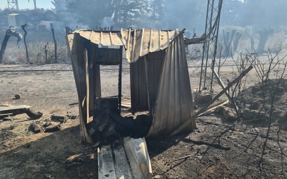 A small shed, warped and charred by a fire. The roof has collapsed in and the surrounding earth is smoking and bare, littered with debris.