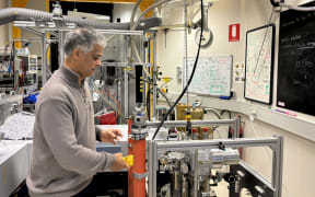 A man in a grey sweater standing side on looks down at a pair of orange cylinders attached to a complex metal frame and scientific equipment with various tubes and pipes. He is a in a lab with an industrial feel.