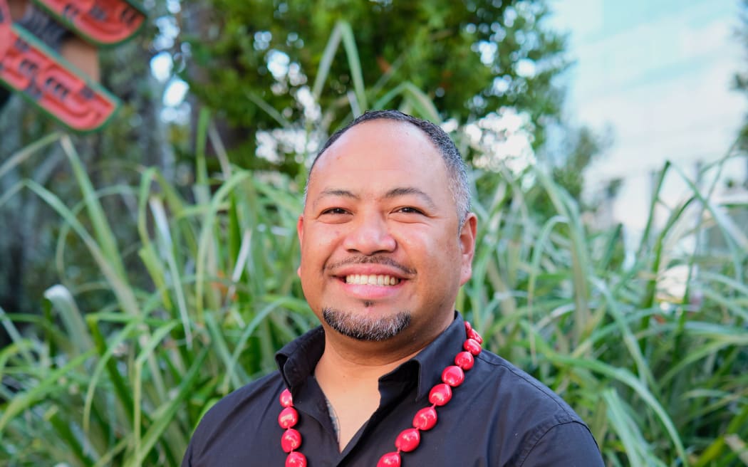 A man wearing a dark shirt standing in front of greenery smiles directly at the camera.
