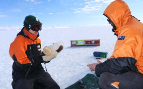 Two people kneeling on a flat white expanse of nows underneath a blue sky. They are both wearing goggles, beanies and orange-and-black warm clothing, and both are holding chunks of ice.