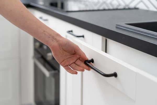 Cropped shot of female hand opening white wooden cupboard door with dark metal handle at kitchen with contemporary furniture.