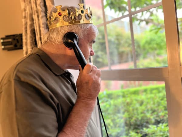 Tom, an elderly man in a brown t-shirt, looks out a window wearing a golden crown and holding an old-fashioned black rotary phone.